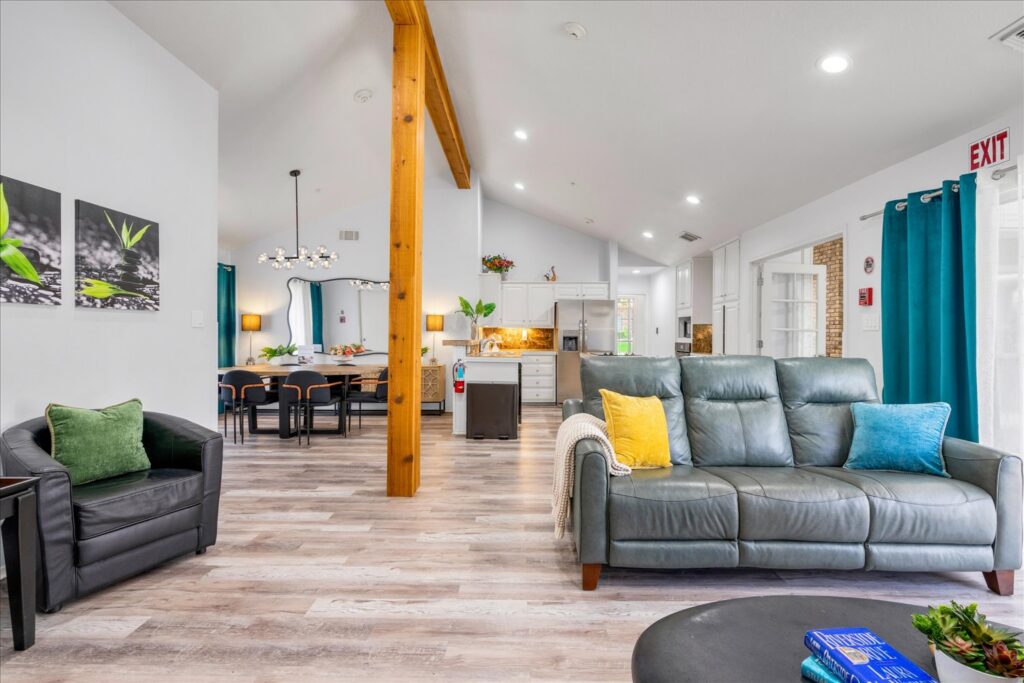 Modern living room with gray sofa, colorful pillows, black armchair, teal curtains, wooden beam, and view into dining area and kitchen.
