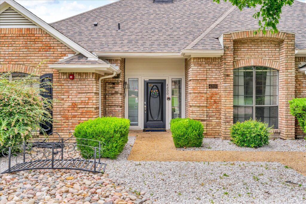 Charming brick house with black front door, decorative glass, large windows with blinds, trimmed shrubs, gravel pathway, and metal fence.