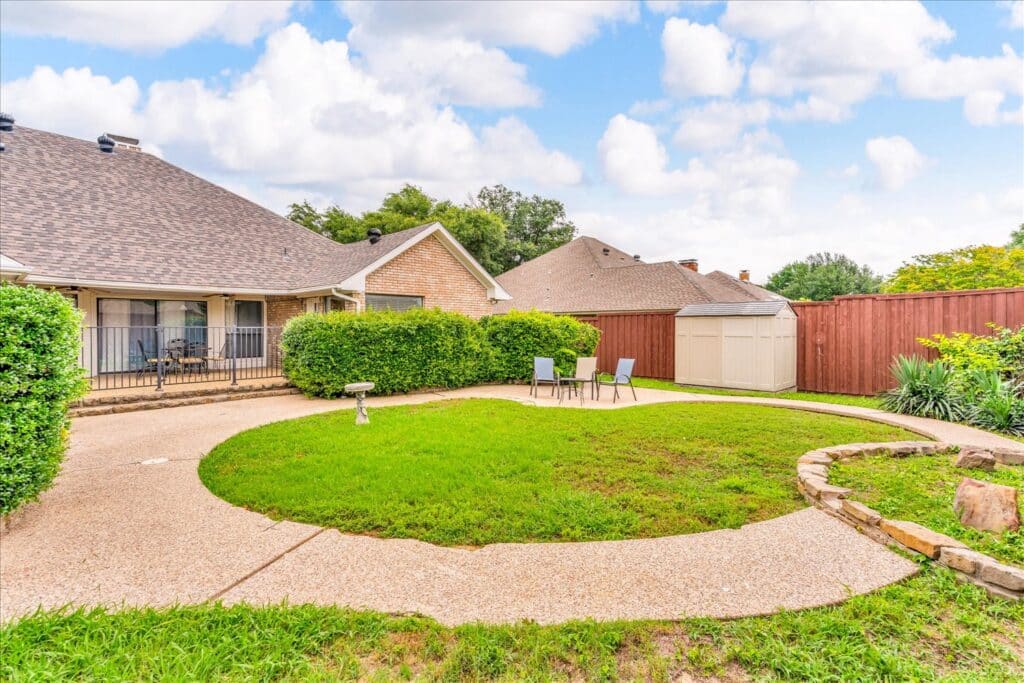 Backyard with circular grassy area, stone pathway, two chairs with table, central birdbath, wooden fence, storage shed, and greenery under a blue sky.