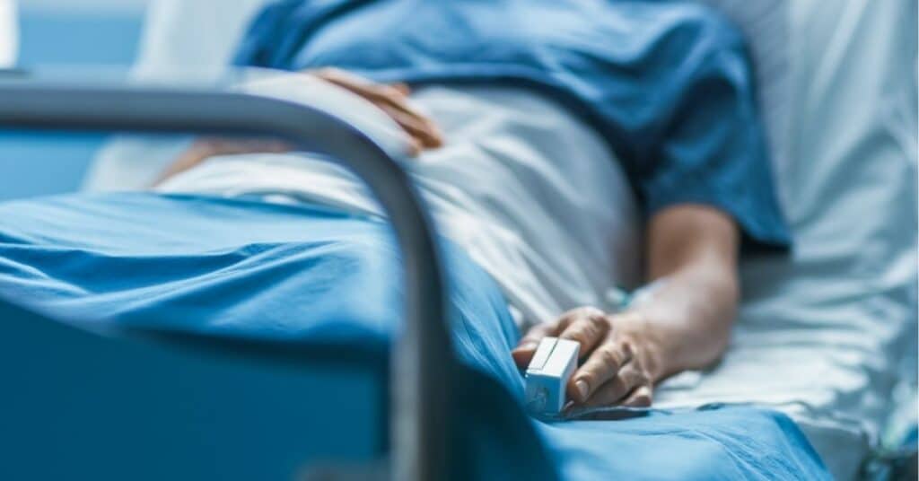 Person in hospital bed wearing blue gown with pulse oximeter, in softly lit medical setting.
