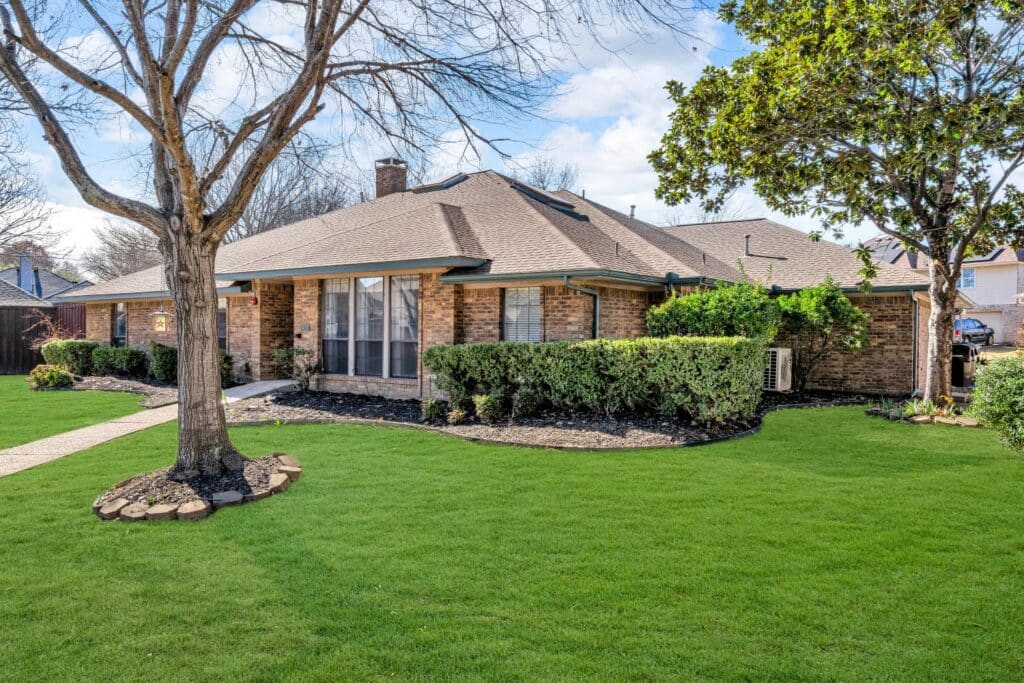 Brick house with sloped roof, green lawn, trimmed bushes, mature trees, and pathway to entrance.