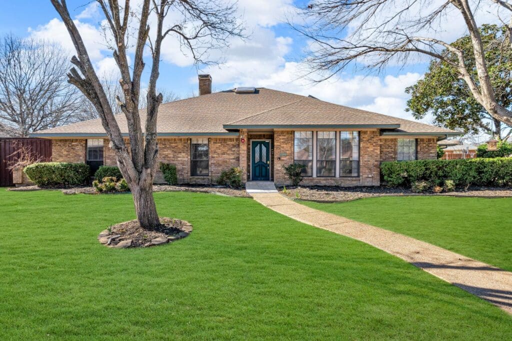 Brick house with sloped roof, green lawn, trimmed bushes, mature trees, and pathway to entrance.