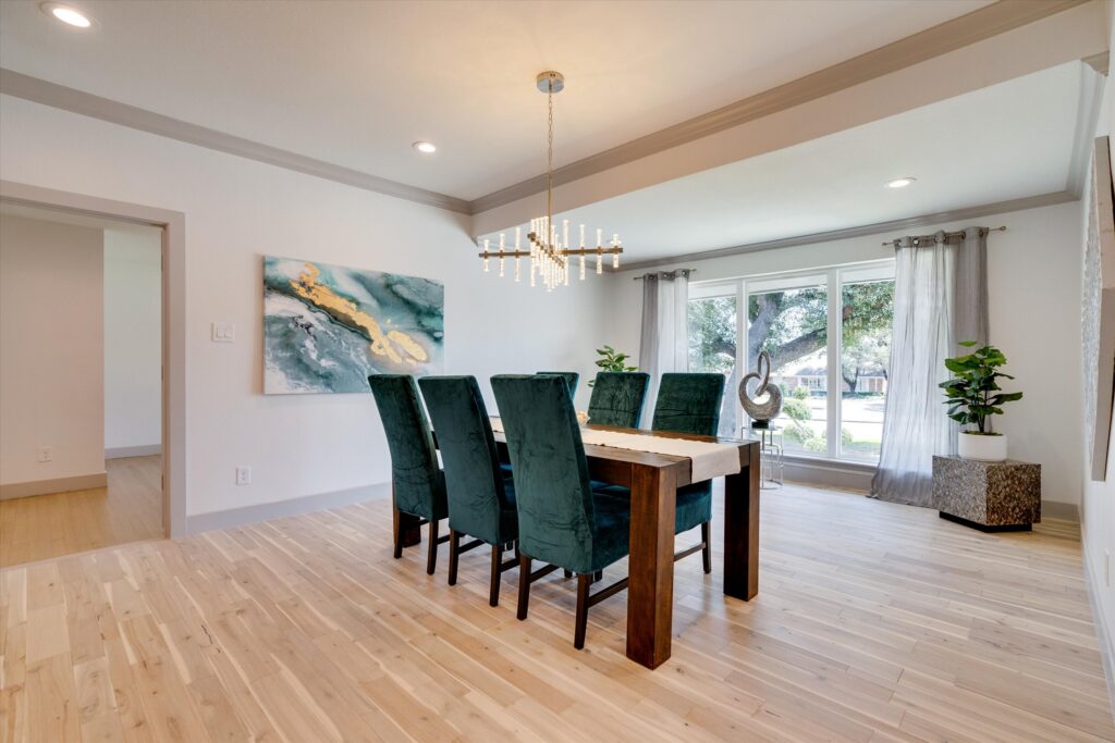 Modern dining room with wooden table, six green velvet chairs, chandelier, abstract painting, and potted plants.