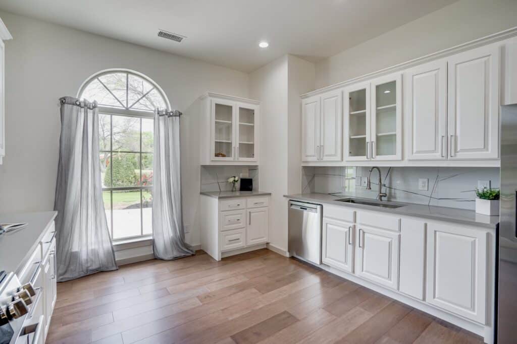 Bright kitchen with white cabinets, gray countertop, stainless steel dishwasher, arched window, and wooden floor.
