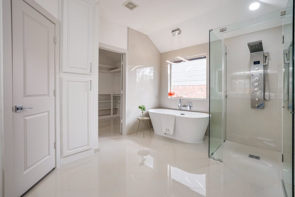 Modern bathroom with freestanding tub by a window, glass shower, walk-in closet, and glossy white tiles.