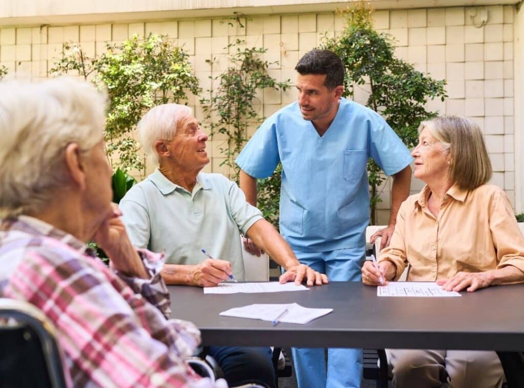 Caregiver in blue scrubs smiling with three seniors at an outdoor table, filling out papers.