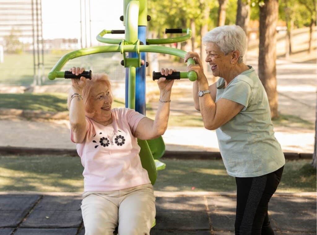 Two older women exercising outdoors, one on green equipment and the other encouraging her, in a sunny, tree-filled area.