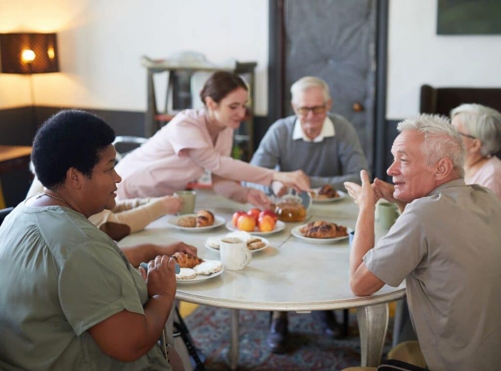 Elderly group sharing pastries and drinks around a table, with a caregiver pouring juice in a cozy room.