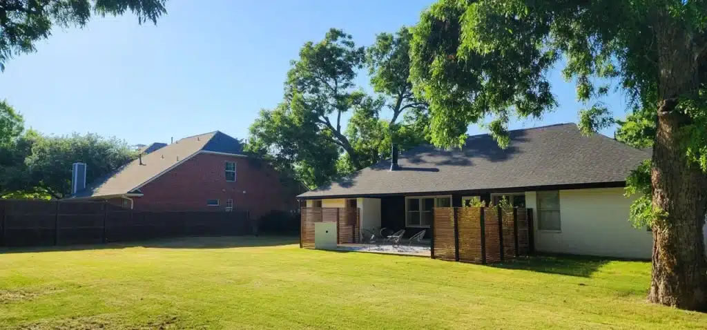 Backyard with mowed lawn, patio seating, wooden privacy screens, and a single-story white house.