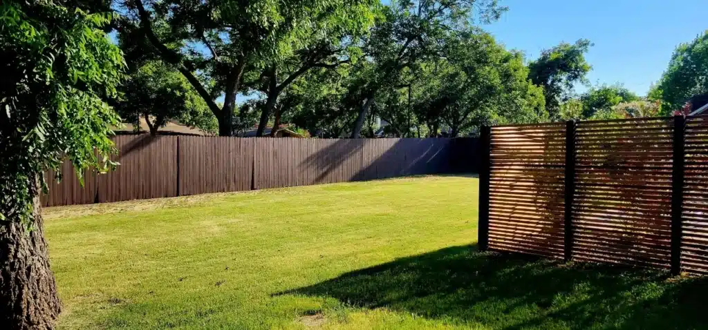 Sunny backyard with trimmed grass, tall wooden fence, and a large tree casting shade.