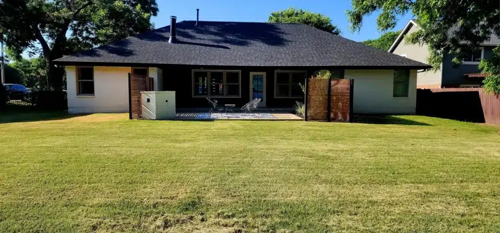 Backyard with mowed lawn, patio seating, wooden privacy screens, and a single-story white house.