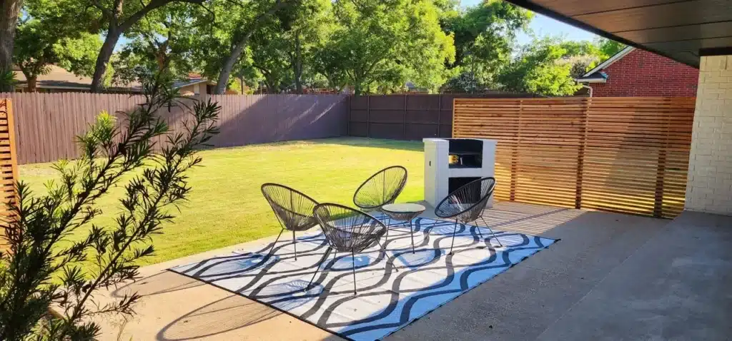 Sunny backyard with green lawn, black patio chairs on patterned rug, and a modern white grill.