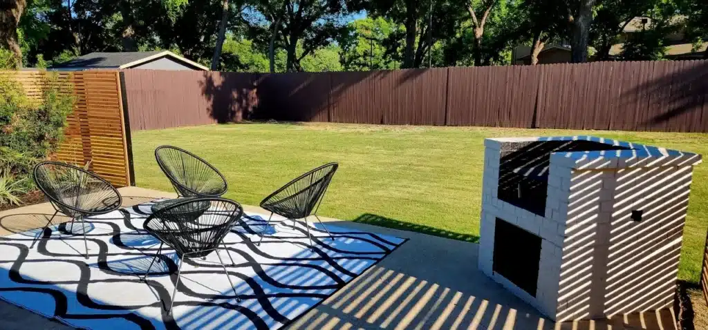 Sunny backyard with green lawn, black patio chairs on patterned rug, and a modern white grill.