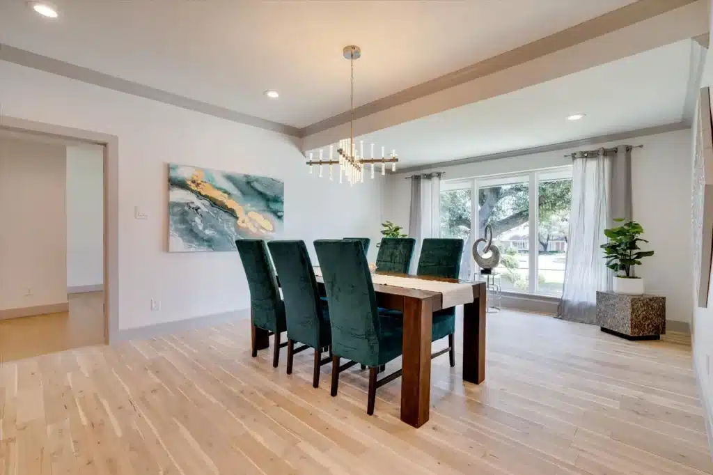 Modern dining room with wooden table, six green velvet chairs, chandelier, abstract painting, and potted plants.