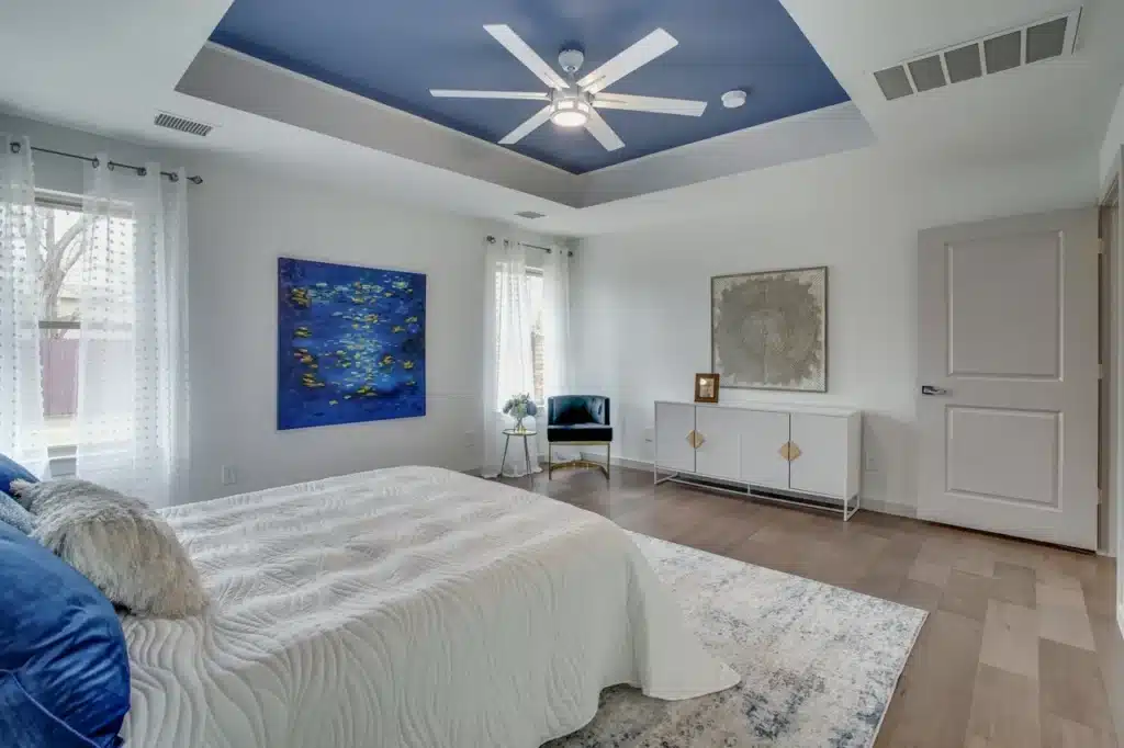 Modern bedroom with white bed, blue accents, abstract painting, ceiling fan, and sheer curtains over hardwood floor.