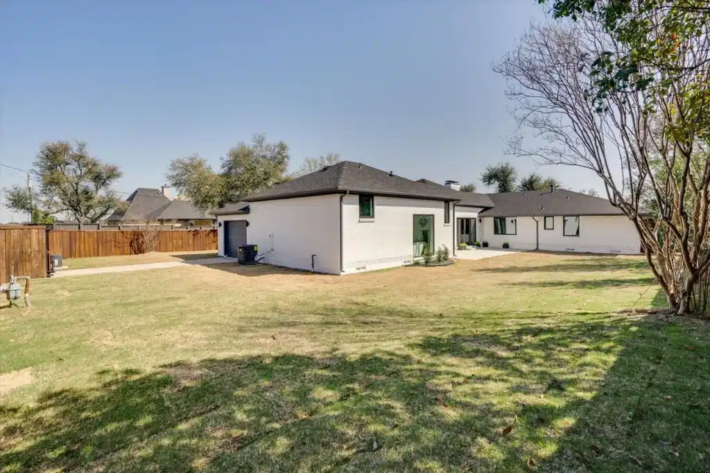 Spacious backyard with modern white house, dark roof, large windows, wooden fence, and trees casting shadows.