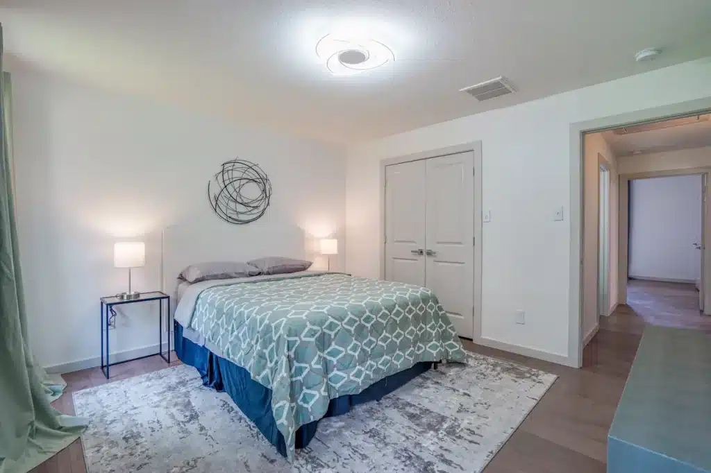 Cozy bedroom with geometric green comforter, bedside tables with lamps, circular wall art, and light wood floor.
