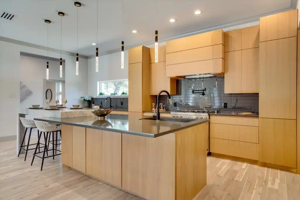 Modern kitchen with light wood cabinets, gray island with black faucet, pendant lights, barstools, and stainless steel stove.