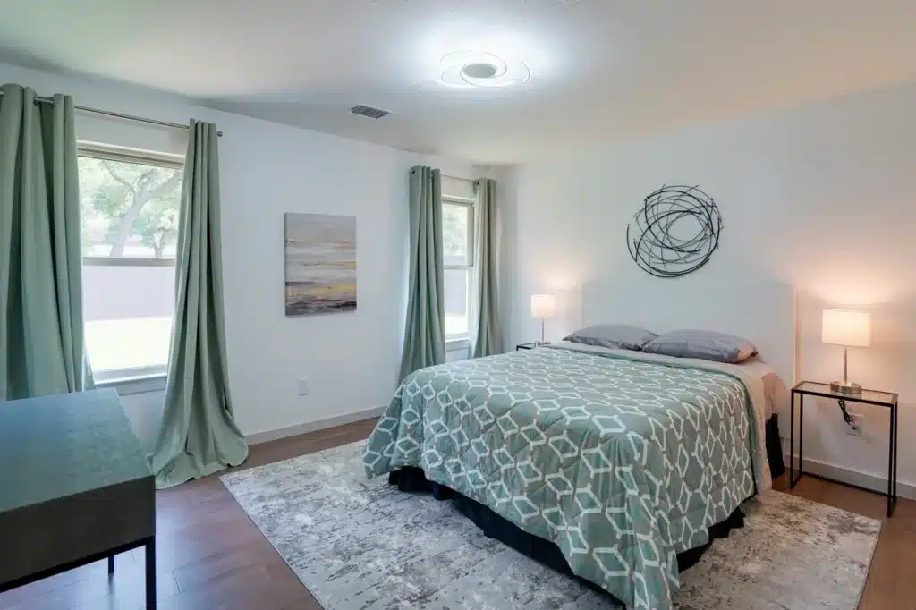 Bright bedroom with green geometric bed, black nightstands with lamps, circular wall sculpture, and wooden floor with gray rug.