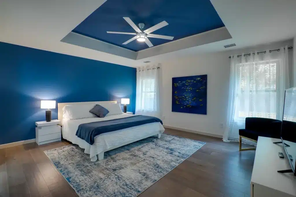 Modern bedroom with white bed, blue accents, abstract painting, ceiling fan, and sheer curtains over hardwood floor.