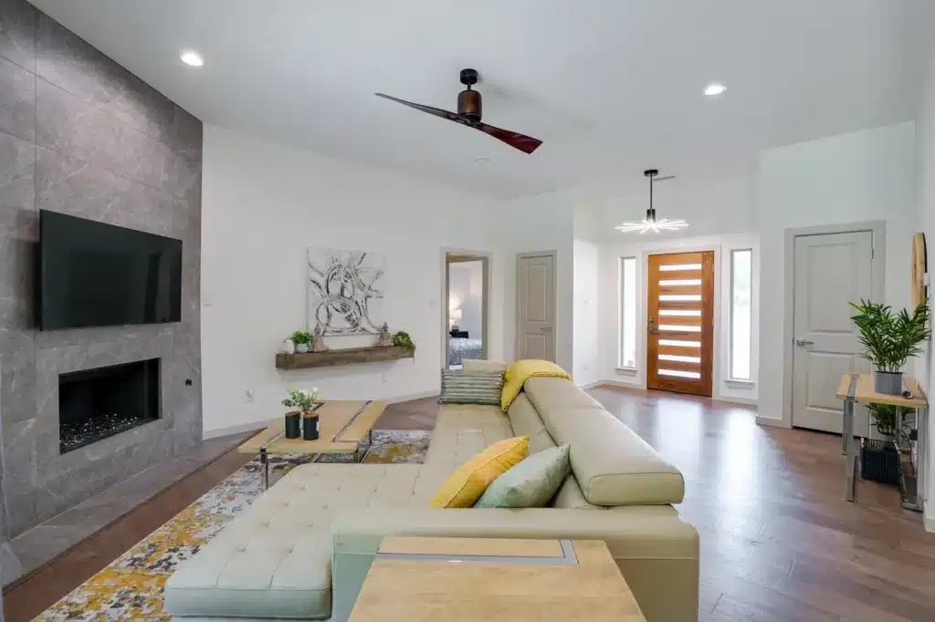Modern living room with beige sectional, colorful cushions, wooden coffee table, stone fireplace, and wall-mounted TV.