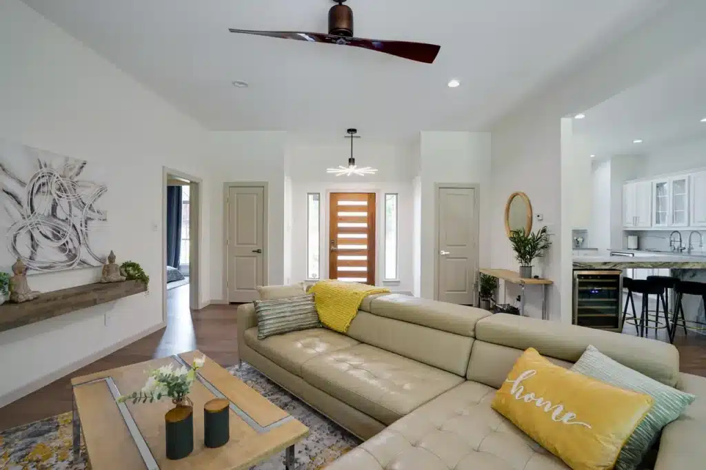 Modern living room with beige sectional, yellow and green cushions, wooden coffee table, ceiling fan, and view of kitchen.