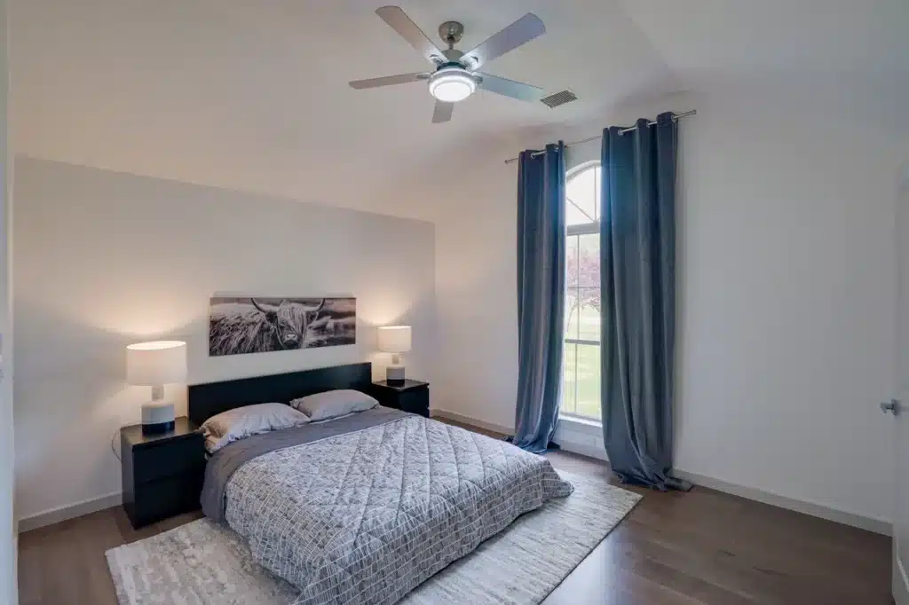 Cozy bedroom with gray bed, black nightstands with lamps, animal photo, dark blue curtains, and ceiling fan.