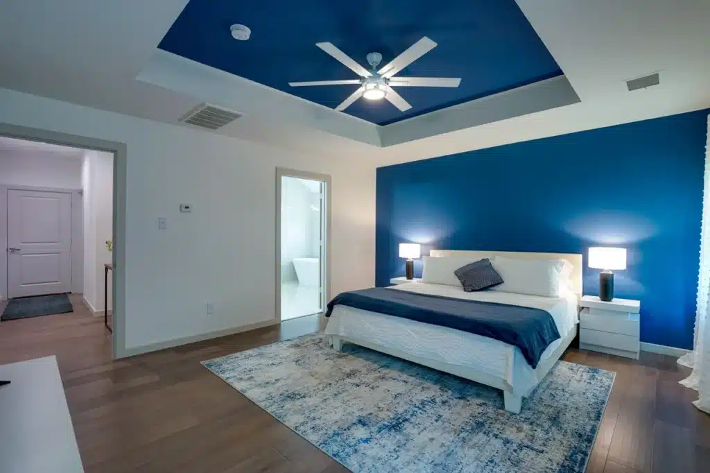 Modern bedroom with white bed, blue accents, abstract painting, ceiling fan, and sheer curtains over hardwood floor.