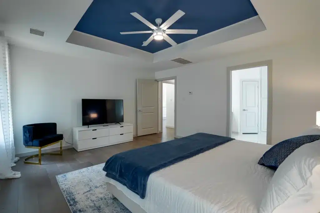 Modern bedroom with white bed and blue accents, white dresser with TV, blue chair, and blue ceiling with ceiling fan.