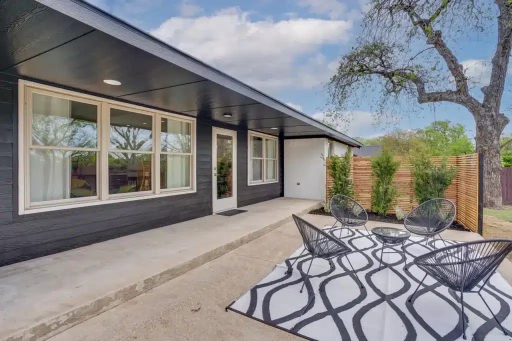 Modern house exterior with black siding, large windows, covered porch, and outdoor seating with a patterned rug.