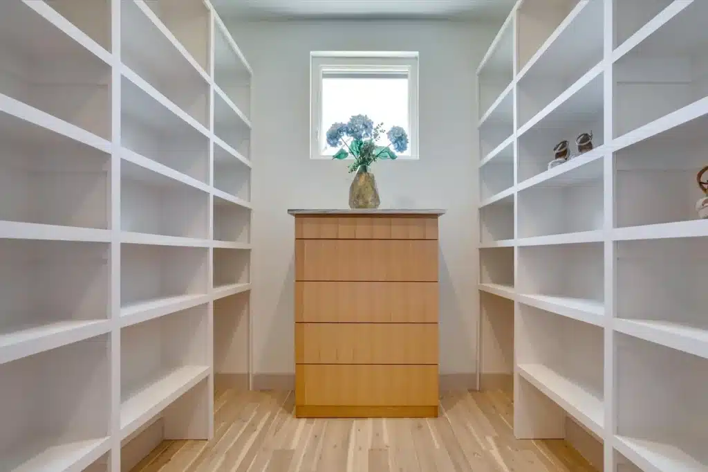 Spacious walk-in closet with white shelving, center dresser with flowers, window, and wooden floor.