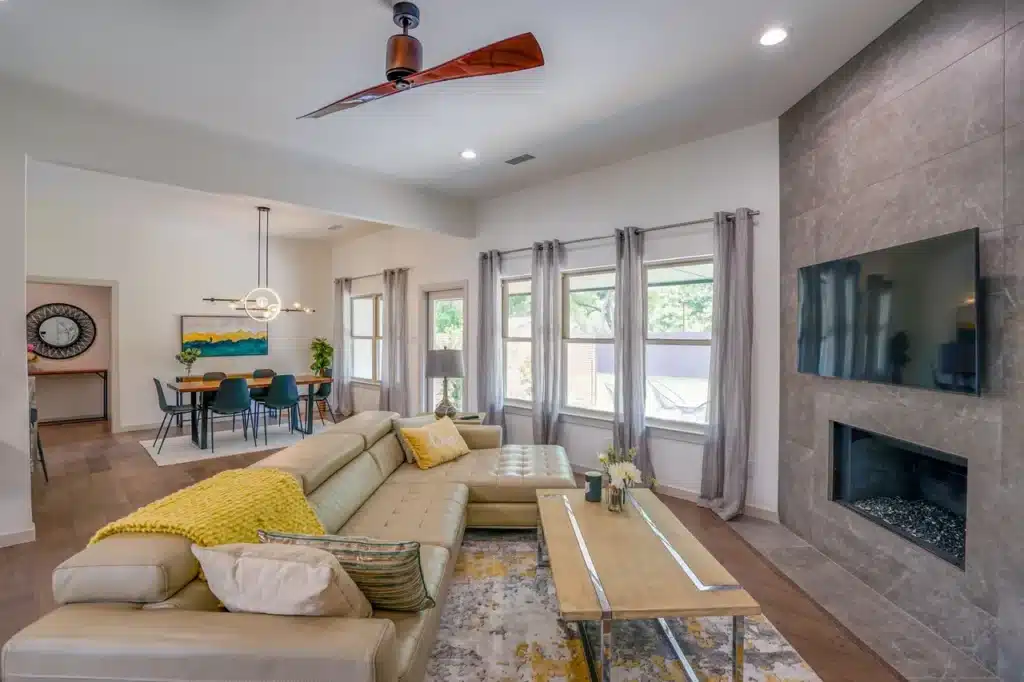 Modern living room with beige sectional, yellow and green cushions, wooden coffee table, ceiling fan, and view of kitchen.