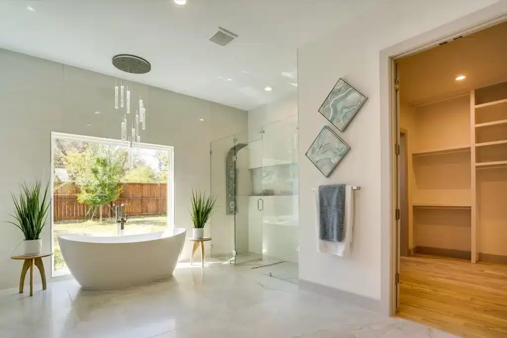Modern bathroom with freestanding tub by window, glass shower, potted plants, towel, and open closet.