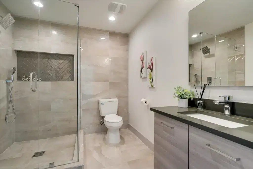 Modern bathroom with glass shower, beige tiles with herringbone accent, white toilet, black vanity, and decor.