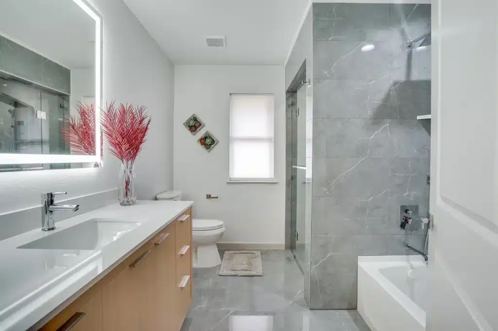 Modern bathroom with light wood vanity, illuminated mirror, glass shower, gray tile floor, and decorative plants.