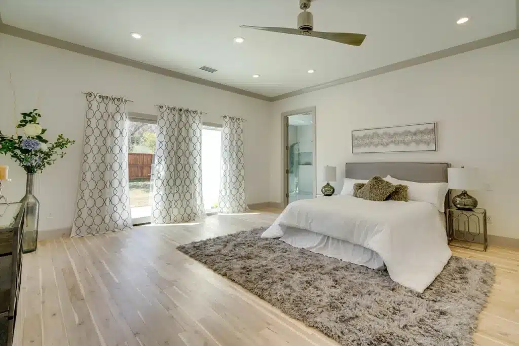 Spacious bedroom with white bed, decorative pillows, gray rug, patterned curtains, ceiling fan, and modern decor.