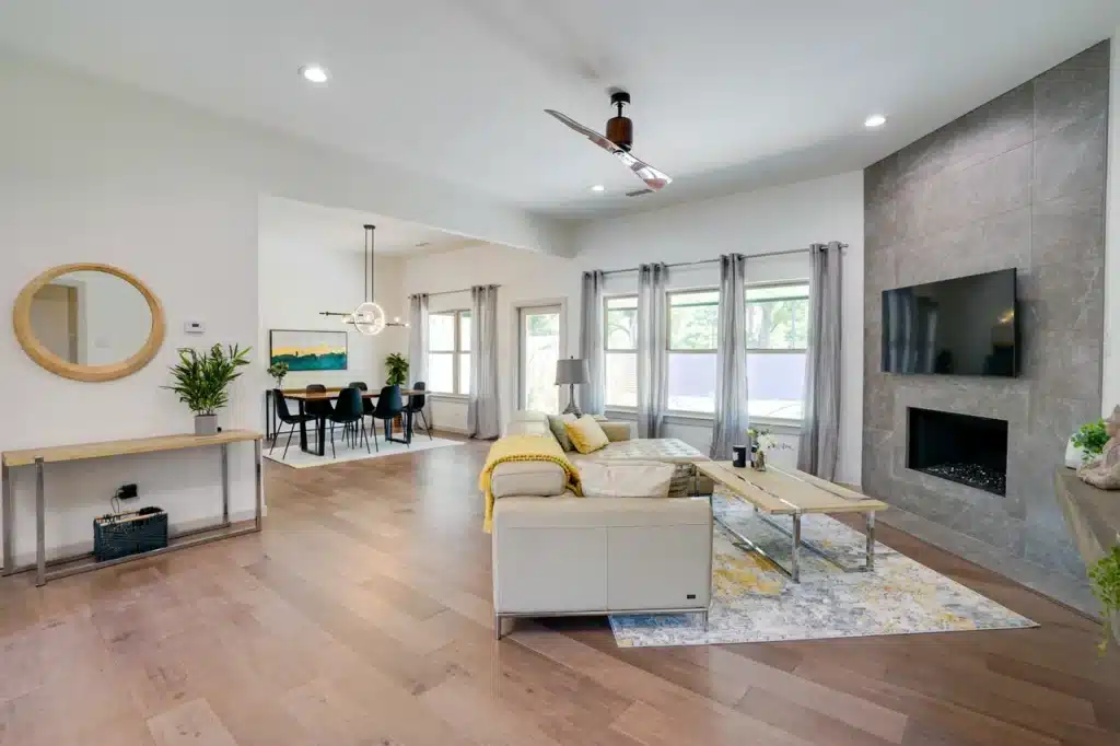 Modern living room with gray fireplace, beige sectional with yellow accents, wooden coffee table, and dining area in the background.