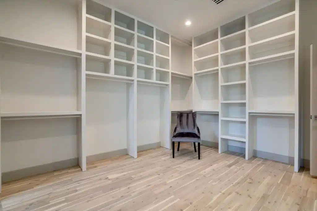 Spacious walk-in closet with white shelves, hanging spaces, and a dark upholstered chair on light wood floor.