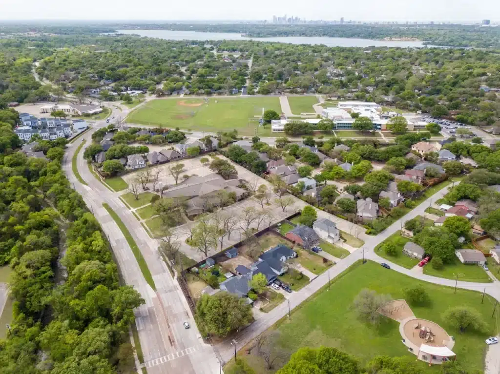 Aerial view of suburban neighborhood with houses, playground, baseball field, winding road, lake, and distant city skyline.