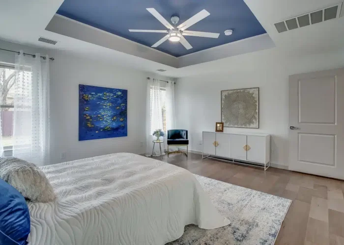 Modern bedroom with white bed, blue accents, abstract painting, ceiling fan, and sheer curtains over hardwood floor.