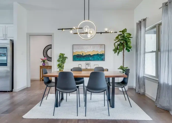 Modern dining room with wooden table, six black chairs, chandelier, abstract painting, and sheer curtains.