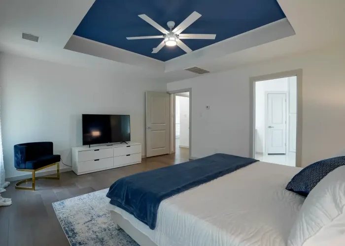 Modern bedroom with white bed and blue accents, white dresser with TV, blue chair, and blue ceiling with ceiling fan.