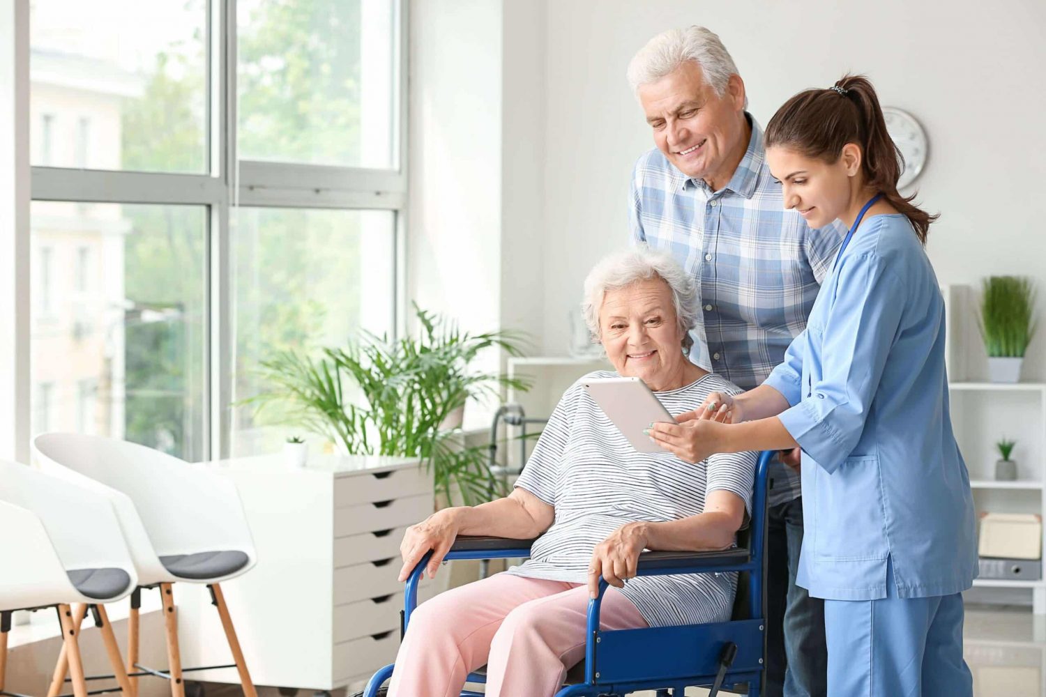 Nurse in blue scrubs showing tablet to elderly woman in wheelchair, with smiling man nearby, in bright room with large windows and plants.